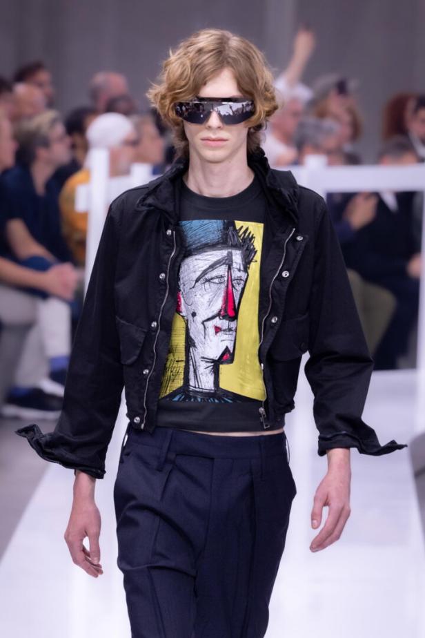 Ein Model mit futuristischer Sonnenbrille und einem T-Shirt mit einem abstrakten Porträt auf einem Laufsteg.