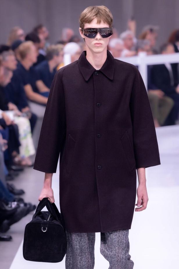 Ein schlanker Mann mit Sonnenbrille und Mantel trägt eine schwarze Tasche.
