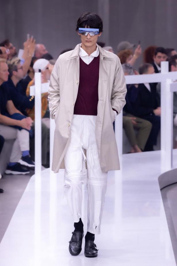 Ein Model mit futuristischer Brille, Trenchcoat und weißen Hosen auf einem Laufsteg.