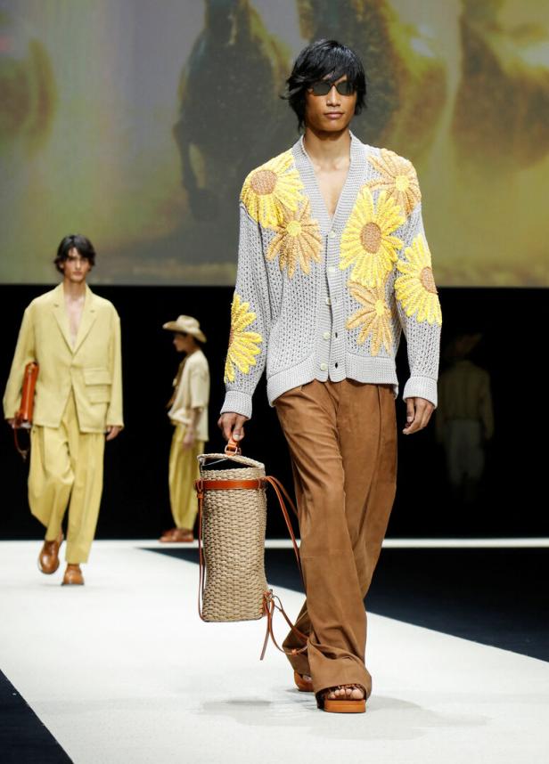 Ein Mann mit Sonnenbrille auf dem Laufsteg, der eine Strickjacke mit Sonnenblumenmuster und eine geflochtene Tasche trägt.