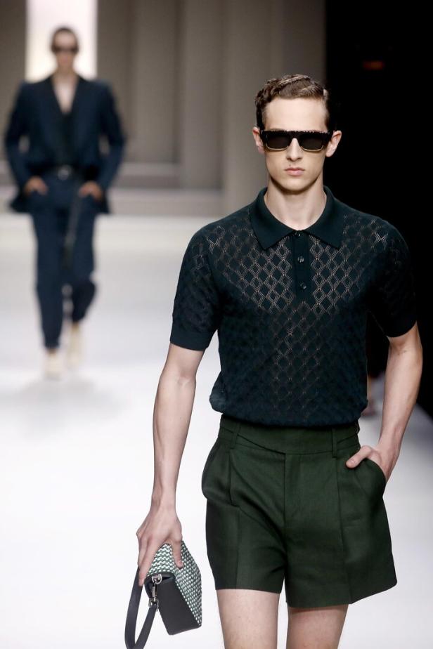 Ein Mann mit Sonnenbrille trägt ein grünes Outfit und eine kleine Tasche auf dem Laufsteg.