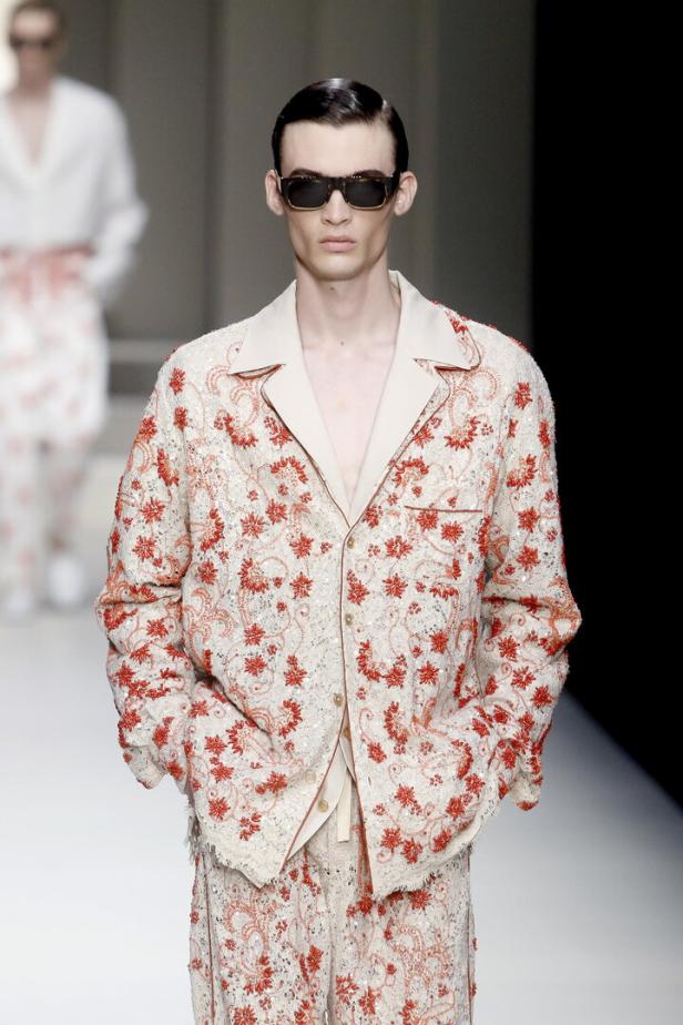Ein Mann mit Sonnenbrille trägt ein weißes Outfit mit roten Blumenmustern auf einem Laufsteg.