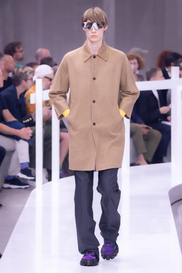 Ein Model mit beigem Mantel, dunkler Hose und auffälligen violetten Schuhen auf einem Laufsteg.