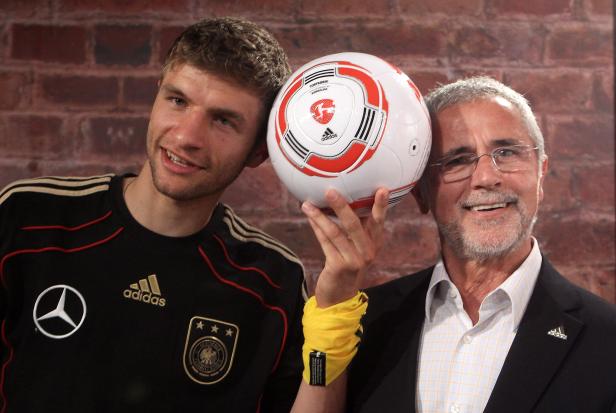 Thomas Müller und ein älterer Mann halten einen Fußball in die Kamera.