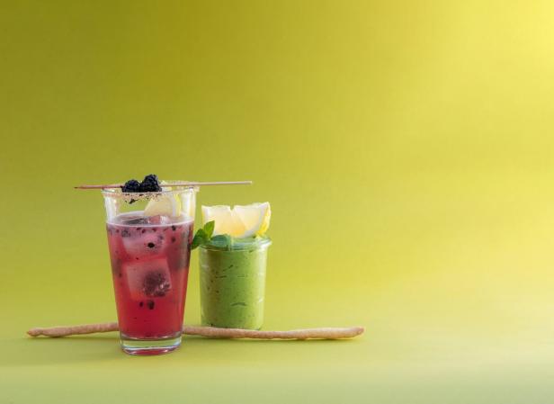 Ein Brombeer-Cocktail mit Eiswürfeln und ein grüner Smoothie auf gelbem Hintergrund.