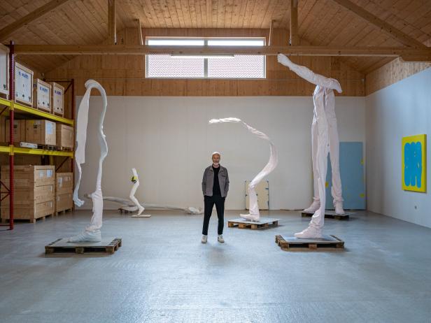 Erwin Wurm steht inmitten seiner skulpturalen Werke in einer Lagerhalle.