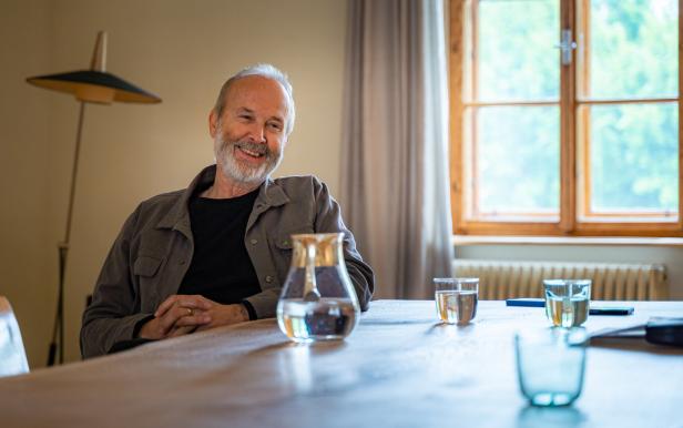 Erwin Wurm sitzt lächelnd an einem Tisch mit Wassergläsern.