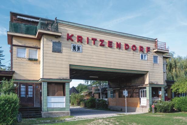 Ein Brückengebäude, das in der Mitte einen Zugang zur Liegewise hat. Es ist schon in die Jahre gekommen und mit Eternitplatten ausgestattet. In roter Schrift steht „KRITZENDORF“ darauf.