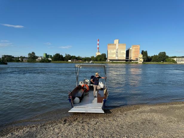 Ein Boot legt am Donauufer in Kritzendorf an. Darauf steht ein Mann mit Hut - im Hintergrund liegt am anderen Ufer Korneuburg.