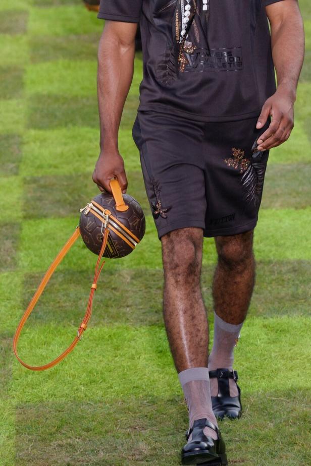 Ein Mann trägt ein Outfit von Louis Vuitton und eine fußballförmige Handtasche.