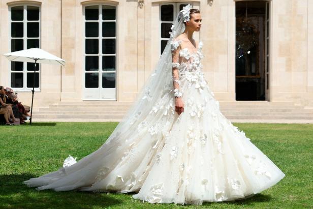 Eine Frau präsentiert ein weißes, blumenverziertes Brautkleid auf einer Modenschau im Freien.