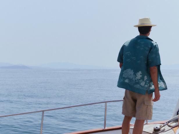 Ein Mann mit Hut und Hemd mit Muster steht auf einem Boot und blickt auf das Meer.