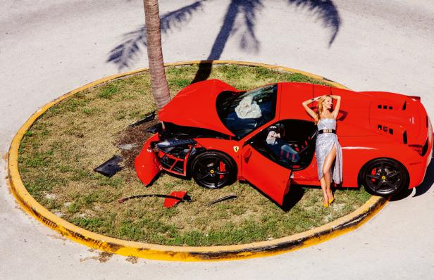 Eine Frau in einem Kleid posiert neben einem roten Ferrari, der gegen eine Palme gekracht ist.