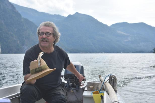 Ein Fischer sitzt in seinem Boot, das auf dem Traunsee fährt. Er hat einen prächtigen Schnurrbart, mit einer Hand lenkt er das Boot, mit der anderen hält er seinen Strohhut.