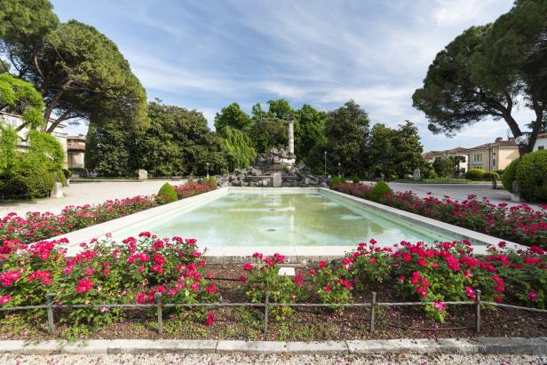 Ein rechteckiger Teich mit einem Säulenmonument im Park ist von roten Rosen umgeben.