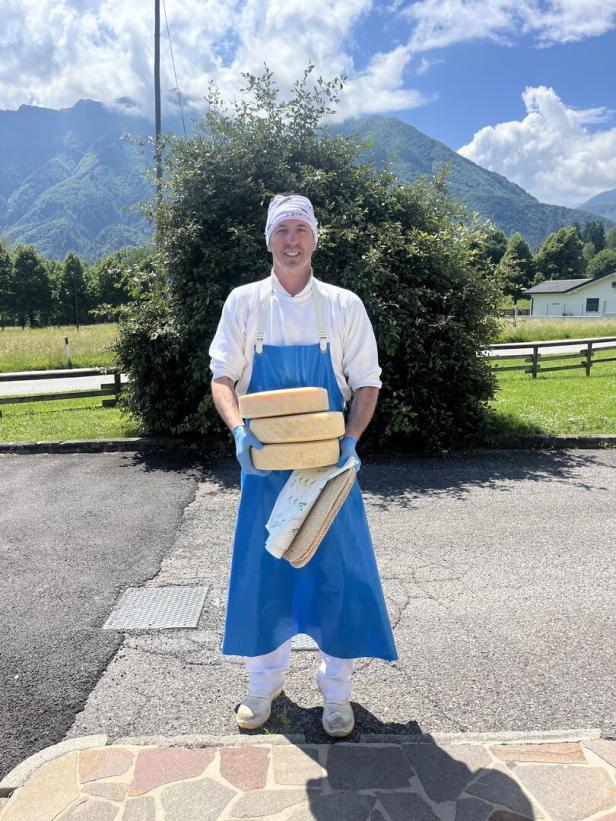Ein Käser in blauer Schürze hält mehrere Laibe Käse vor einer Bergkulisse.