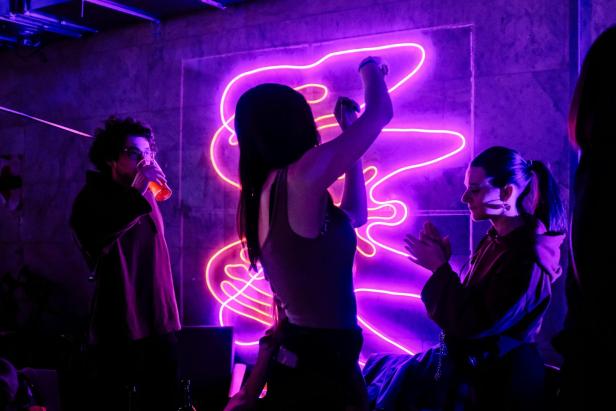 Menschen tanzen und unterhalten sich vor einer leuchtenden Neoninstallation in einem Club.
