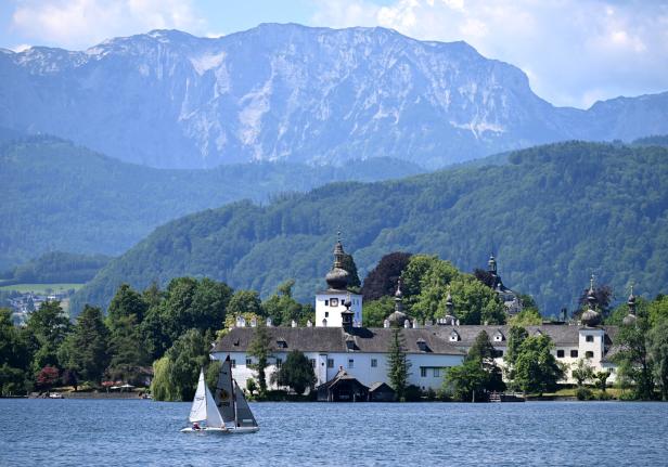 Ein Segelboot fährt vor dem Schloss Orth im Traunsee. Im Hintergrund bauen sich große Berge auf.