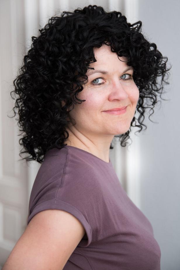 Eine Frau mit lockigem, dunklem Haar und einem lila T-Shirt lächelt in die Kamera.