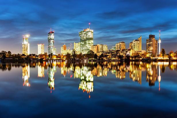 Die beleuchtete Skyline der Wiener Donauplatte in der Nacht. Davor liegt die Alte Donau, in der sich die Hochhäuser spiegeln.