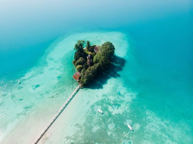 Luftaufnahme vom Attersee: ein Steg führt zu einer kleinen, grünen Insel. Rundherum ist türkisblaues Wasser - Boote schwimmen darauf.