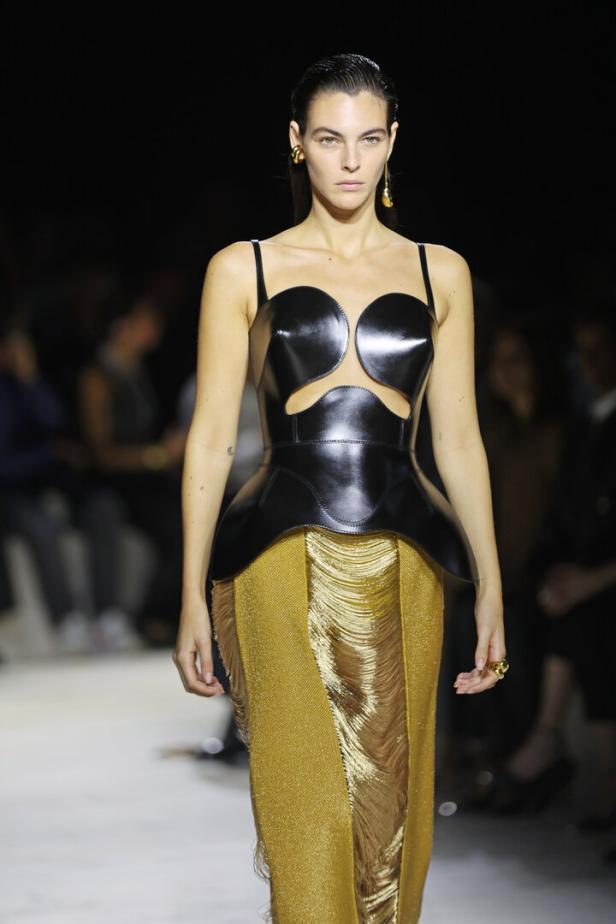 Eine Frau auf dem Laufsteg, gekleidet in ein schwarzes Lackoberteil und einen goldenen Fransenrock.