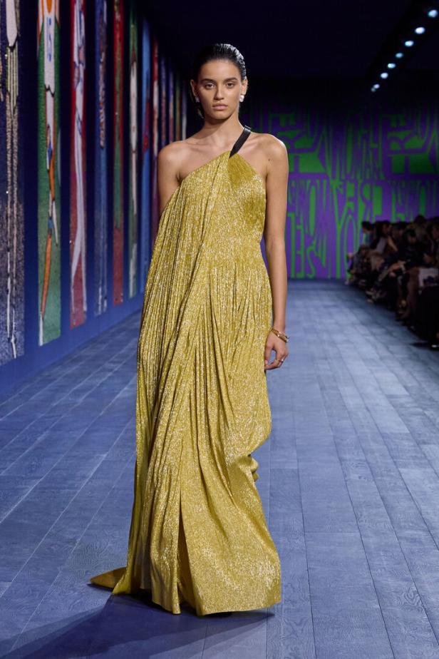 Ein Model präsentiert ein goldenes, plissiertes Kleid auf einem Laufsteg.