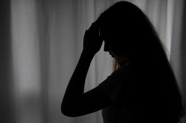 Die Silhouette einer Frau mit gesenktem Kopf und einer Hand an der Stirn vor einem Vorhang.