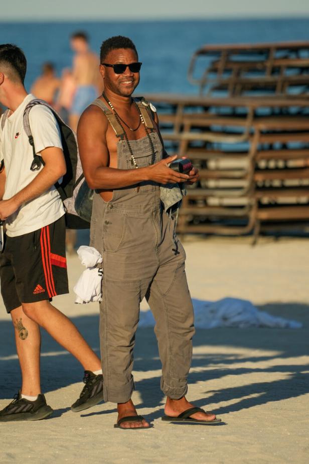 Ein Mann mit Sonnenbrille und Latzhose steht am Strand.