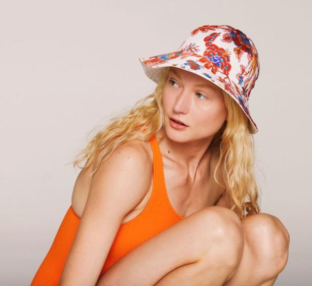 Eine Frau mit blonden Haaren trägt einen orangefarbenen Badeanzug und einen floral gemusterten Hut.