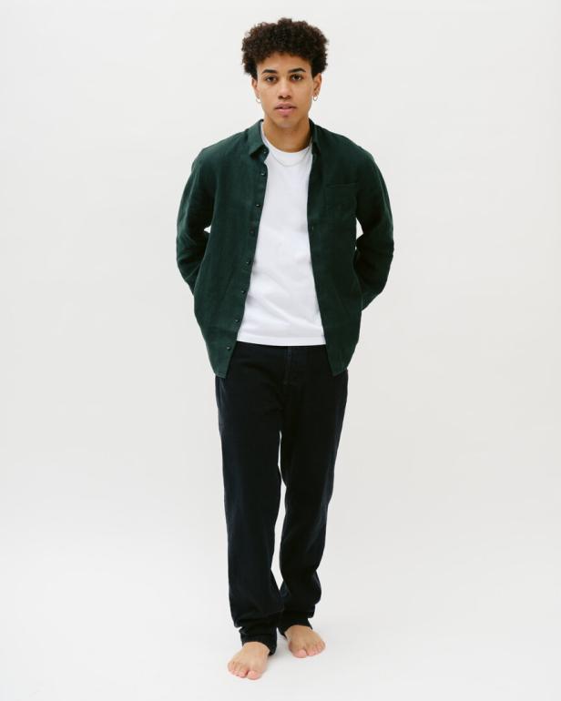 Ein barfüßiger Mann mit lockigem Haar trägt eine grüne Jacke, ein weißes T-Shirt und dunkle Jeans.