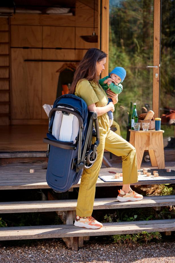 Eine Frau mit Kinderwagen über der Schulter trägt ein Baby auf einer Holztreppe.