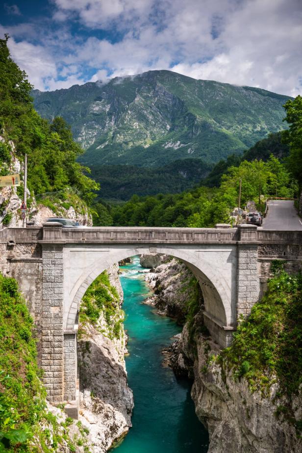 Eine Steinbrücke überspannt einen türkisfarbenen Fluss in einer grünen Landschaft.