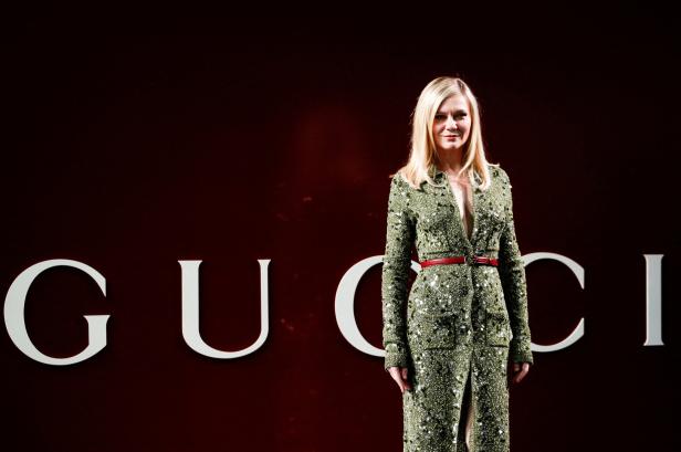 Kirsten Dunst in einem grünen Gucci-Kleid mit rotem Gürtel vor einem Gucci-Logo.