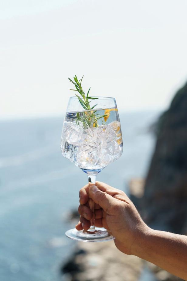 Eine Hand hält ein Glas mit einem Gin Tonic, Eis und Rosmarin vor einem Meerblick.