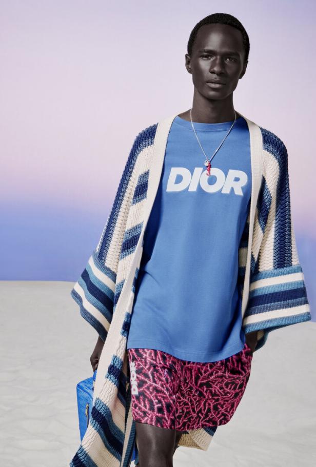 Ein dunkelhäutiger Mann trägt ein blaues Dior-T-Shirt, Shorts mit Korallenmuster und eine Strickjacke.