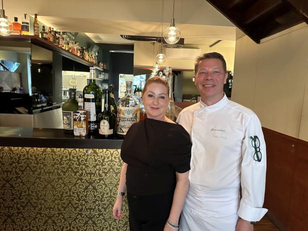 Ein Koch in Kochjacke und eine Frau stehen vor einer Bar mit Spirituosenflaschen.