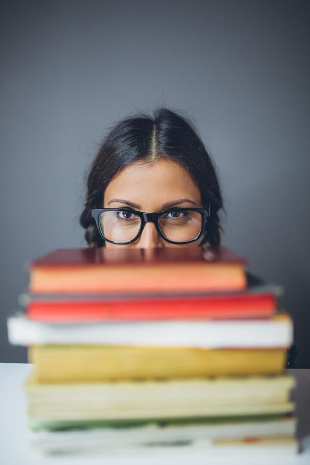 Eine Frau mit Brille schaut über einen Stapel Bücher.