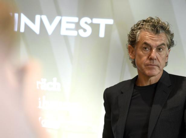 Ein Mann mit grauem, lockigem Haar steht vor einer Leinwand mit dem Wort „Invest“.