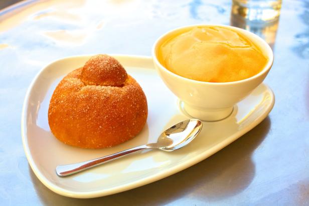 Ein Brioche mit Zucker und eine Schale mit gelber Creme auf einem Teller.