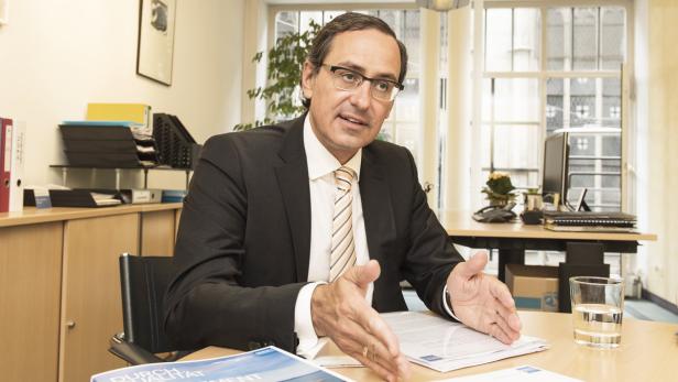 Ein Mann mit Brille und Anzug sitzt an einem Schreibtisch in einem Büro.