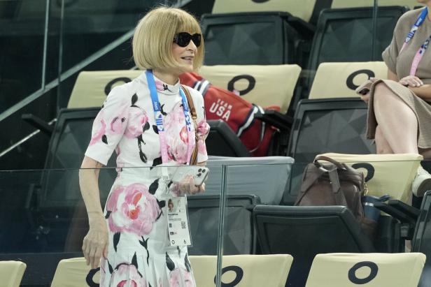 Anna Wintour, in einem floralen Kleid und mit Sonnenbrille, sitzt auf der Tribüne.