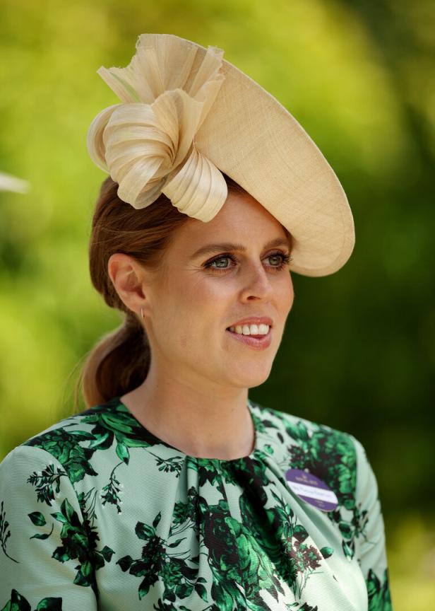 Prinzessin Beatrice mit beigem, kunstvoll verziertem Hut und grün gemustertem Kleid vor unscharfem, grünem Hintergrund.