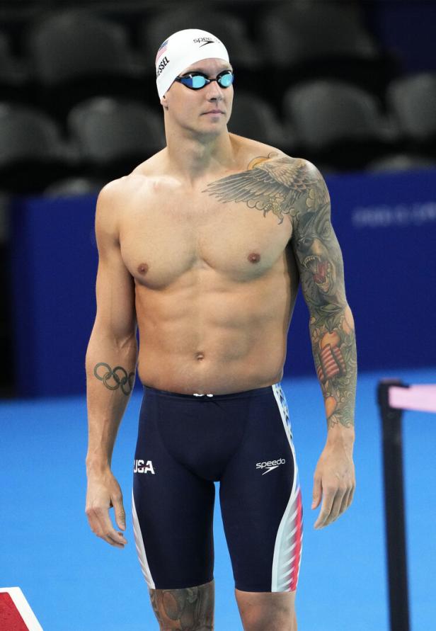Ein tätowierter Schwimmer mit Badekappe und Schwimmbrille steht am Beckenrand.