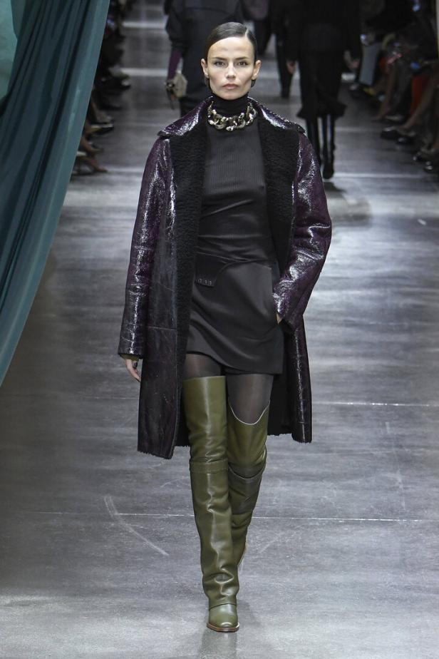 Eine Frau in einem schwarzen Kleid, einem glänzenden Mantel und grünen Stiefeln auf einem Laufsteg.