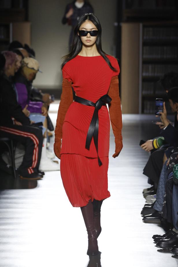 Eine Frau mit Sonnenbrille präsentiert ein rotes Kleid mit braunen Ärmeln und einem schwarzen Gürtel auf dem Laufsteg.