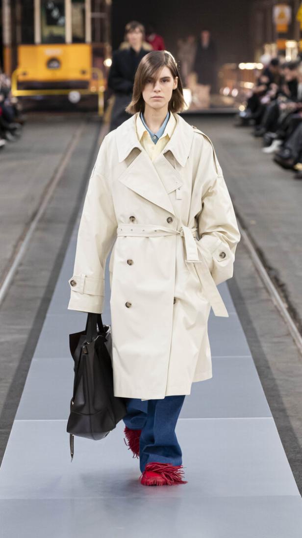 Eine Frau in einem Trenchcoat mit einer schwarzen Tasche auf einem Laufsteg.
