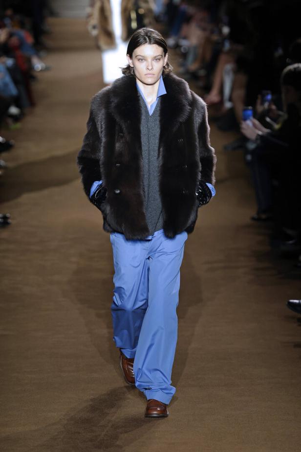 Ein Model in einem braunen Pelzmantel und blauen Hosen auf einem Laufsteg.