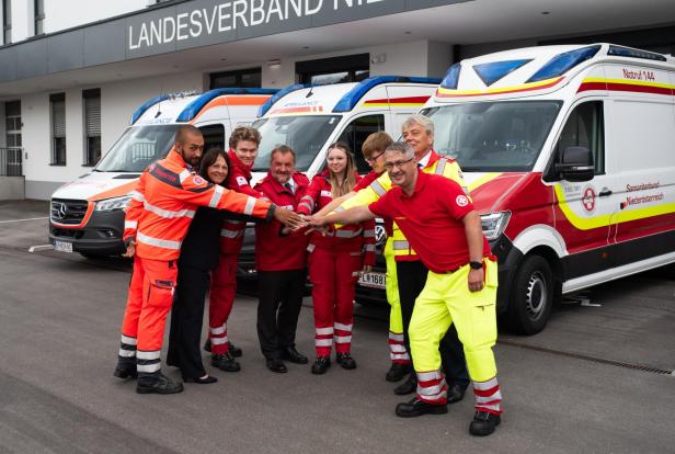 Mehrere Mitglieder der NÖ Rettungsorganisationen stehen vor drei Rettungsfahrzeugen und halten die Hände zusammen.