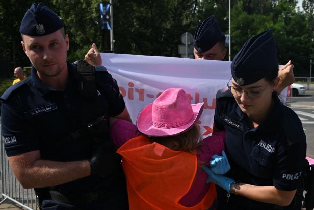 Polizisten führen eine Demonstrantin mit pinkem Hut und orangefarbener Weste ab.